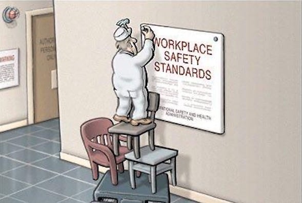 A man nailing a "workplace safety standards" sign while standing on very unsafely stacked chairs